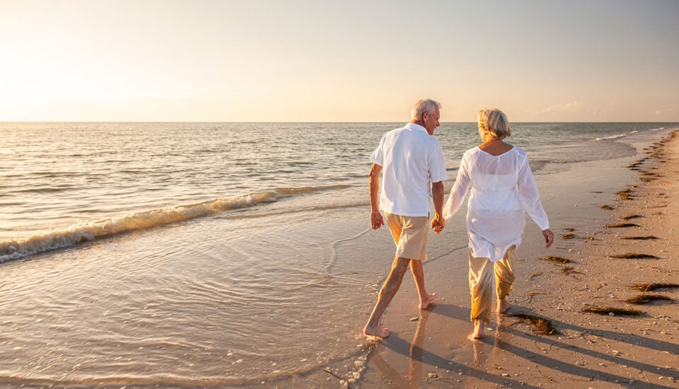 An older couple walking barefoot along the shoreline at sunset